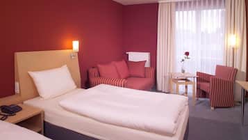 A hotel room with a large bed, a smaller bed, a sofa, a chair, a table, and a vase with a flower.