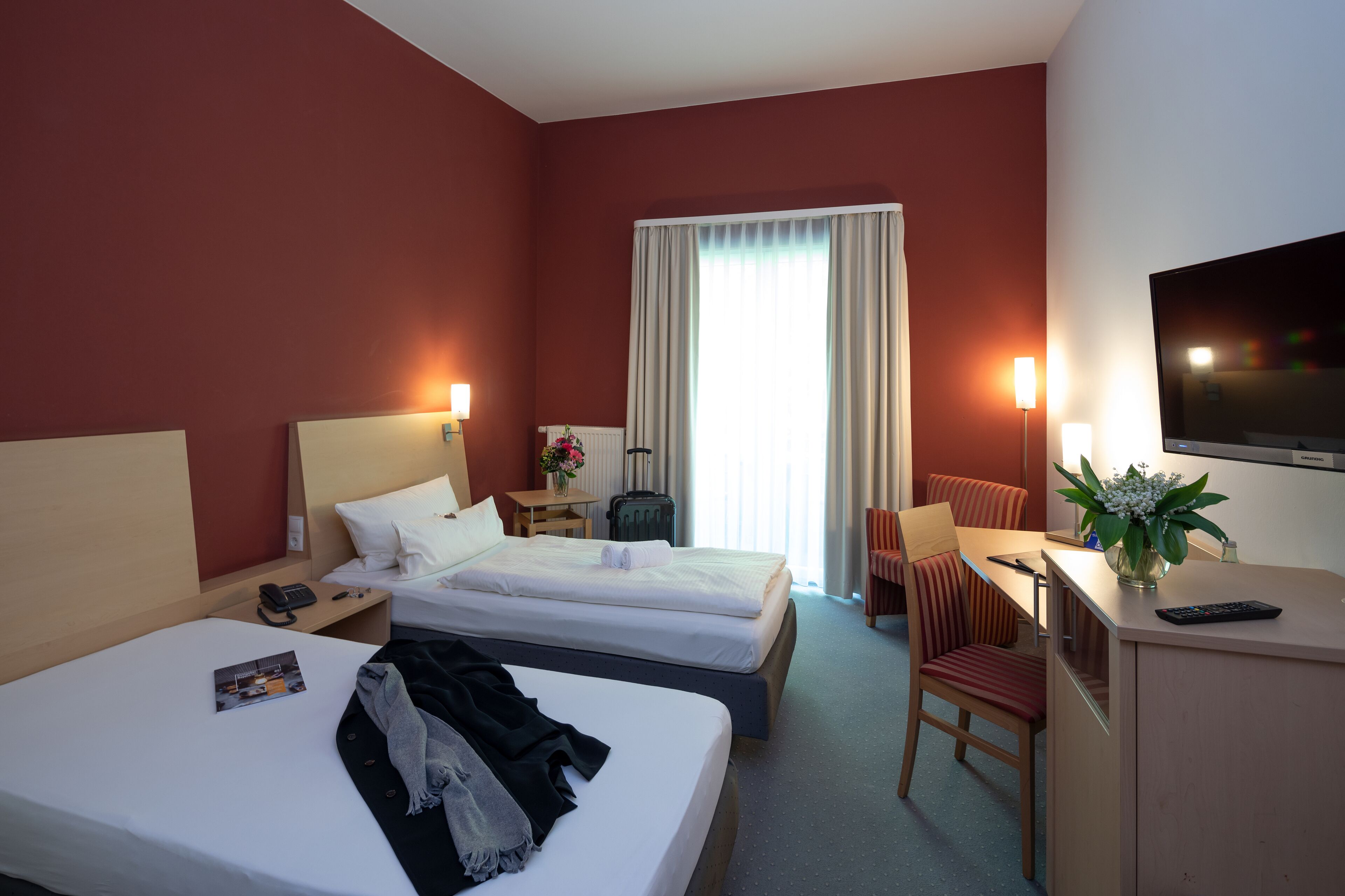 A hotel room with two beds, a desk, a TV, and a window with curtains.