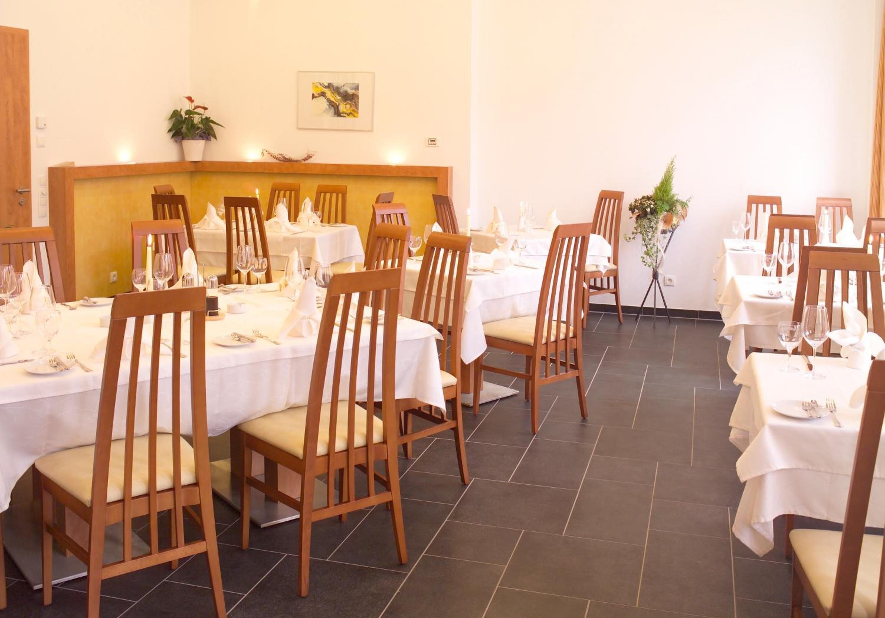 Un coin repas avec des chaises et des tables en bois, préparé pour un repas, agrémenté d’un arrangement floral central et d’un tableau au mur.