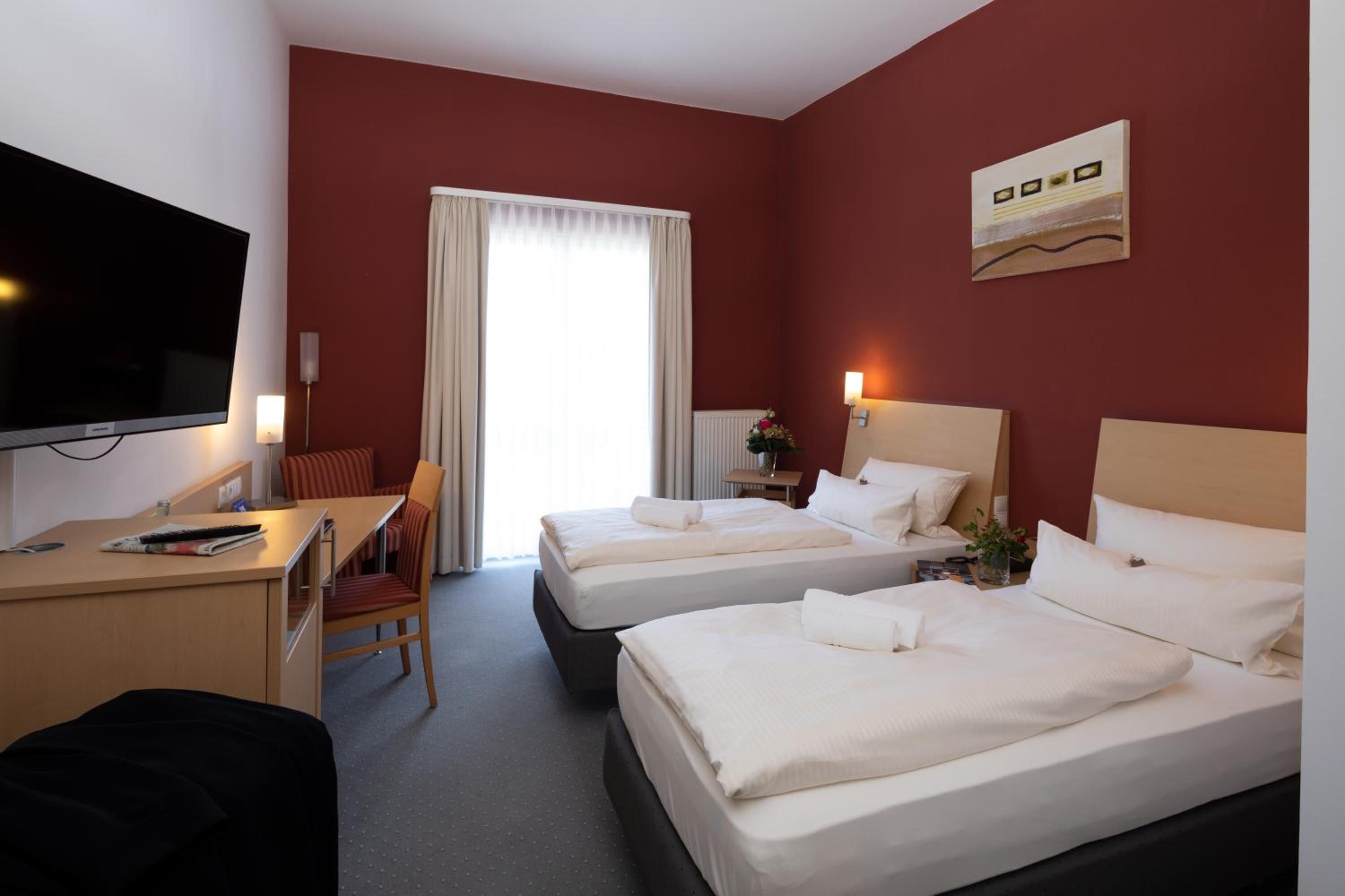 Habitación de hotel con dos camas, un escritorio con silla, televisor y ventana con cortinas.