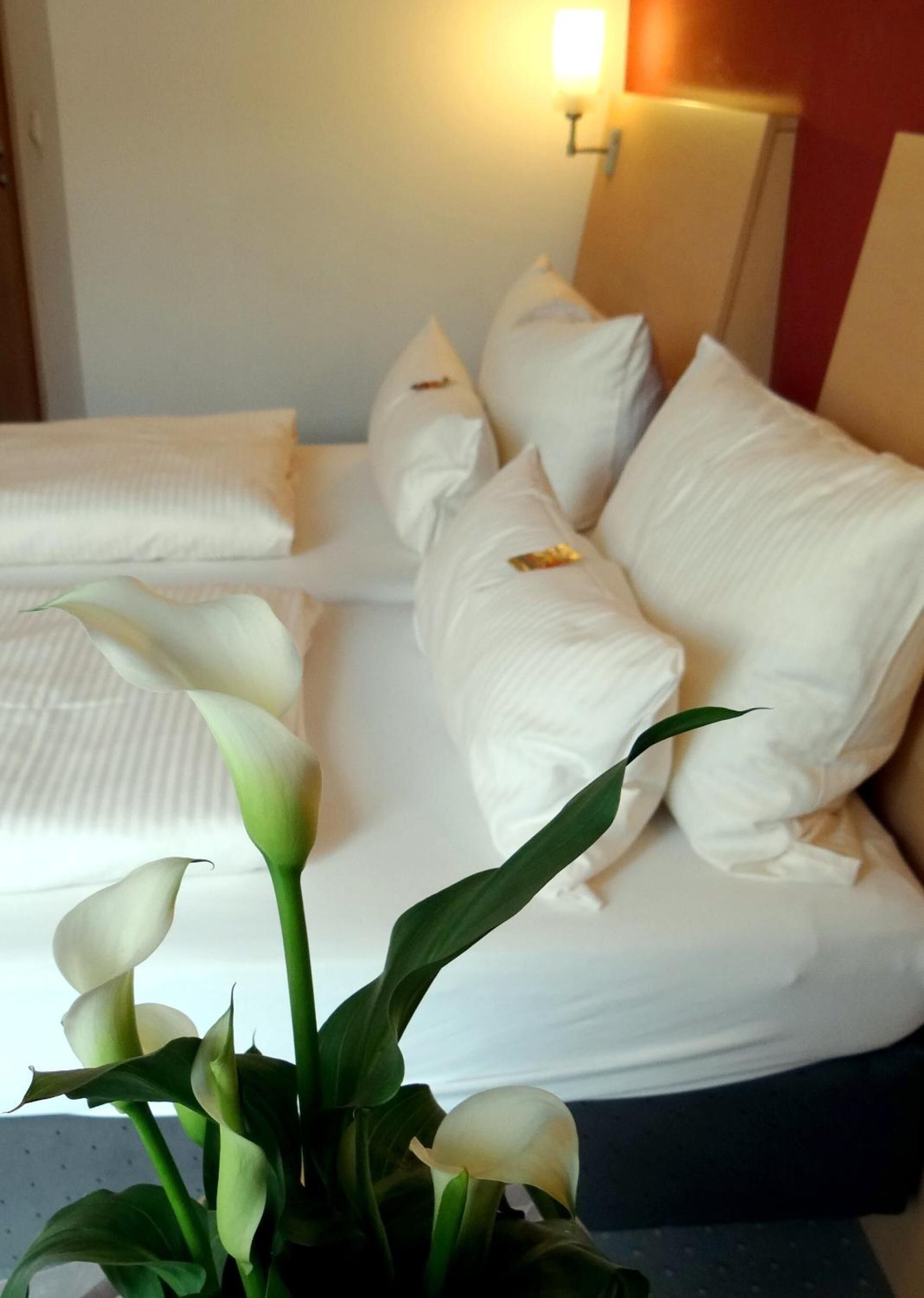 Una habitación de hotel con una cama bien hecha, ropa de cama blanca y un jarrón con flores blancas.