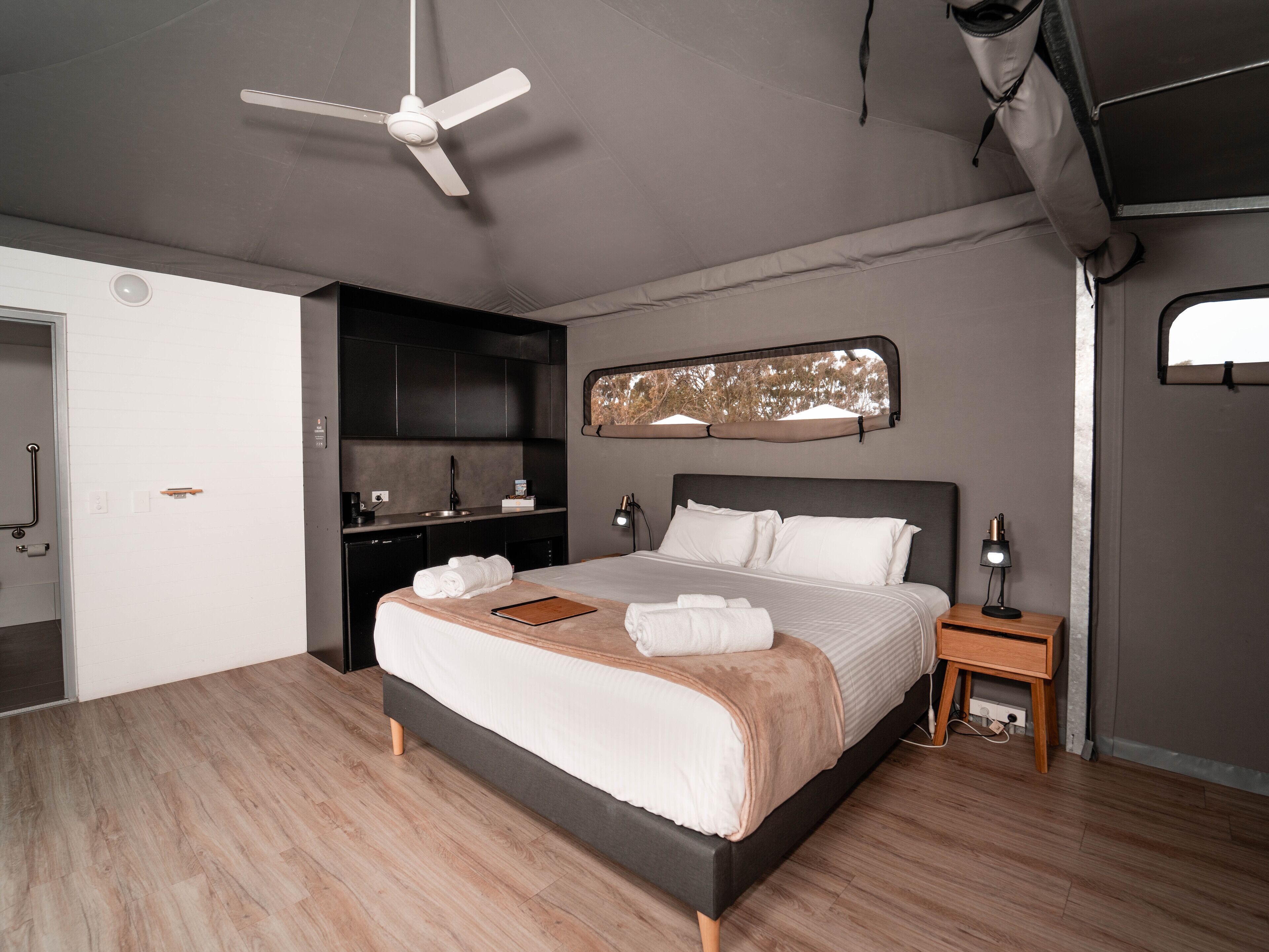 A modern bedroom with a bed, bedside tables, a ceiling fan, a built-in kitchen area, and a large window.