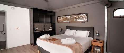 A modern bedroom with a bed, bedside tables, a ceiling fan, a built-in kitchen area, and a large window.