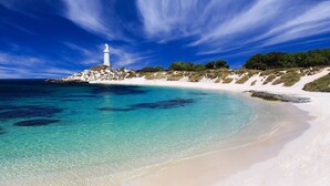A coastal scene with a lighthouse on a rocky outcrop, clear turquoise waters, and a sandy beach.