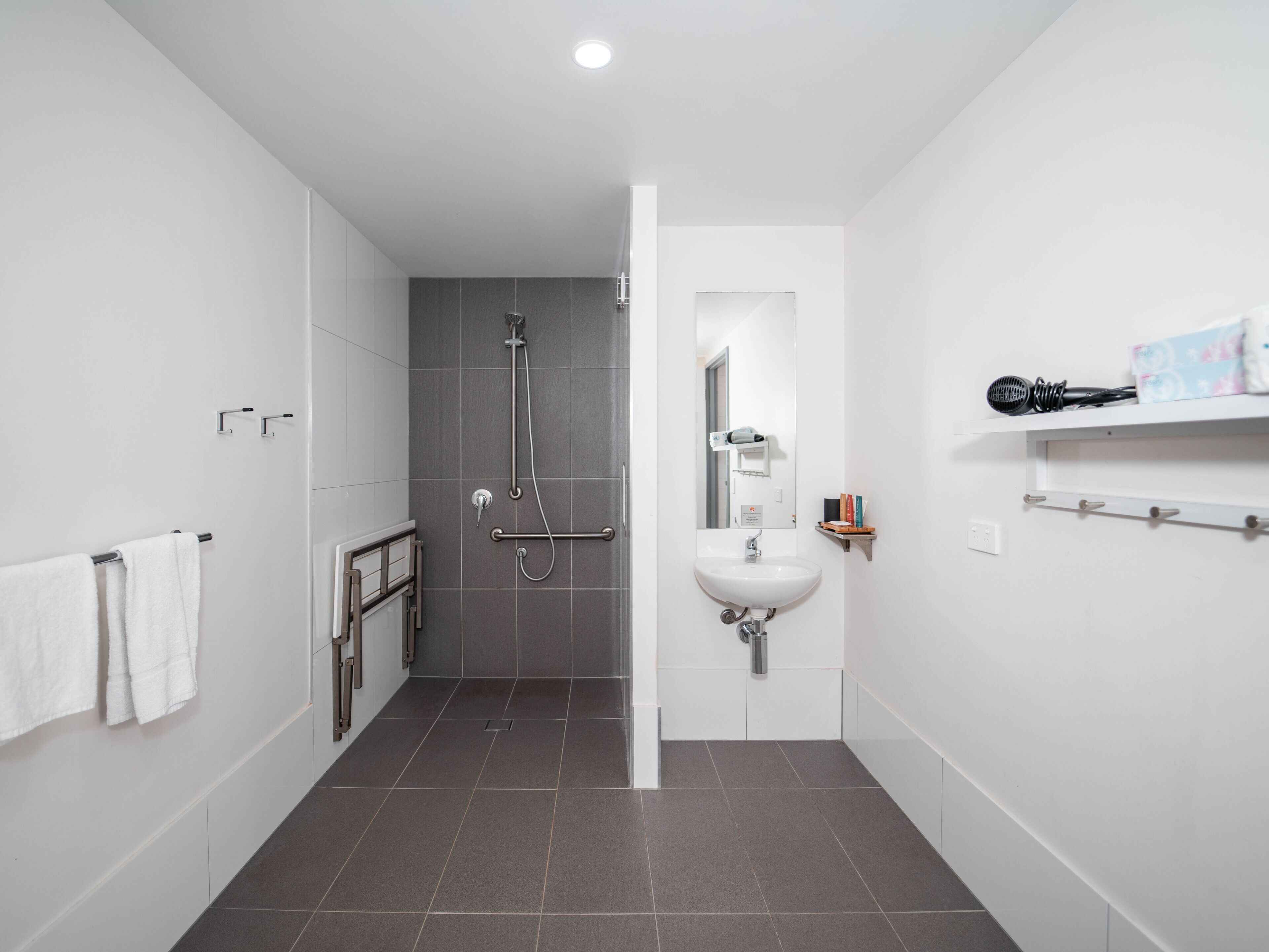A modern bathroom with a shower, a sink, and a mirror. There is a towel rack and a shelf with toiletries.