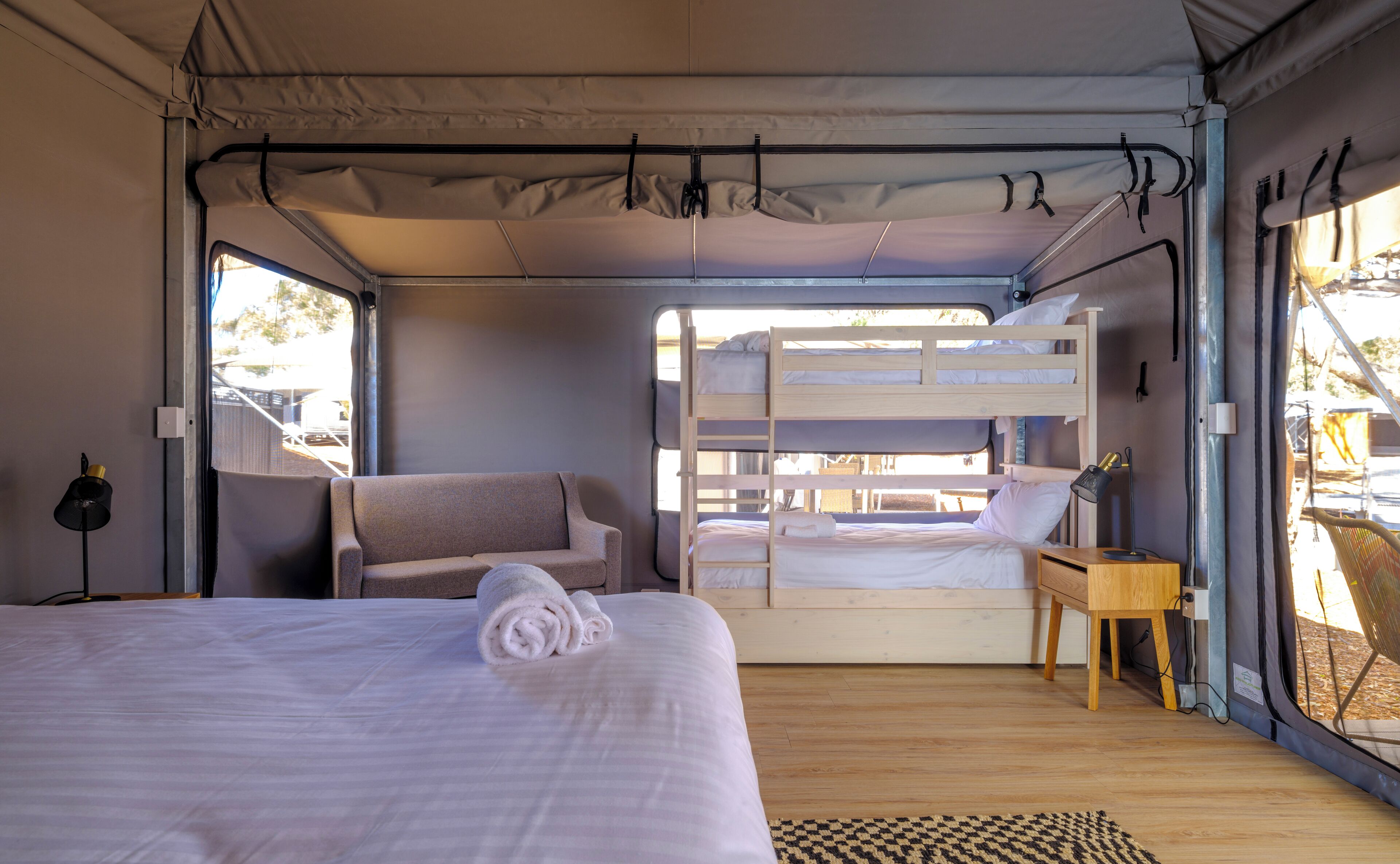 A tent-style room with a bunk bed, a single bed, a chair, a small table, and a lamp.