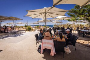An outdoor dining area with tables and chairs under large umbrellas, overlooking a beach.