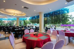 A spacious indoor dining area with round tables, white chairs, and a view of greenery through large windows.