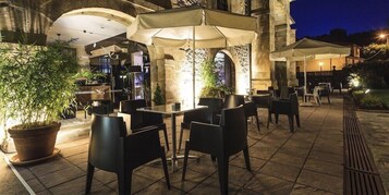 Un patio al aire libre con mesas y sillas bajo sombrillas, rodeado de muros de piedra y macetas con plantas.