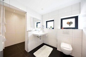 A modern bathroom with white tiles, a white sink, a mirror, a toilet, and a shower area. - Hotel Restaurant Sengscheider Hof (Sankt Ingbert)