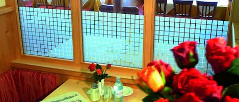 Un comedor con una mesa dispuesta para dos personas, un periódico, una botella y un florero con flores.