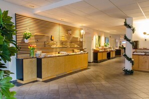 Un espace d’accueil d’hôtel avec un comptoir de réception en bois, un écran mural et un sol carrelé.