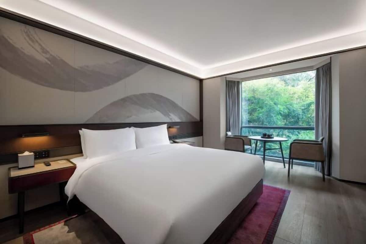 Una habitación de hotel moderna con una cama grande, mesita de noche, lámpara y vistas a zonas verdes por una ventana.