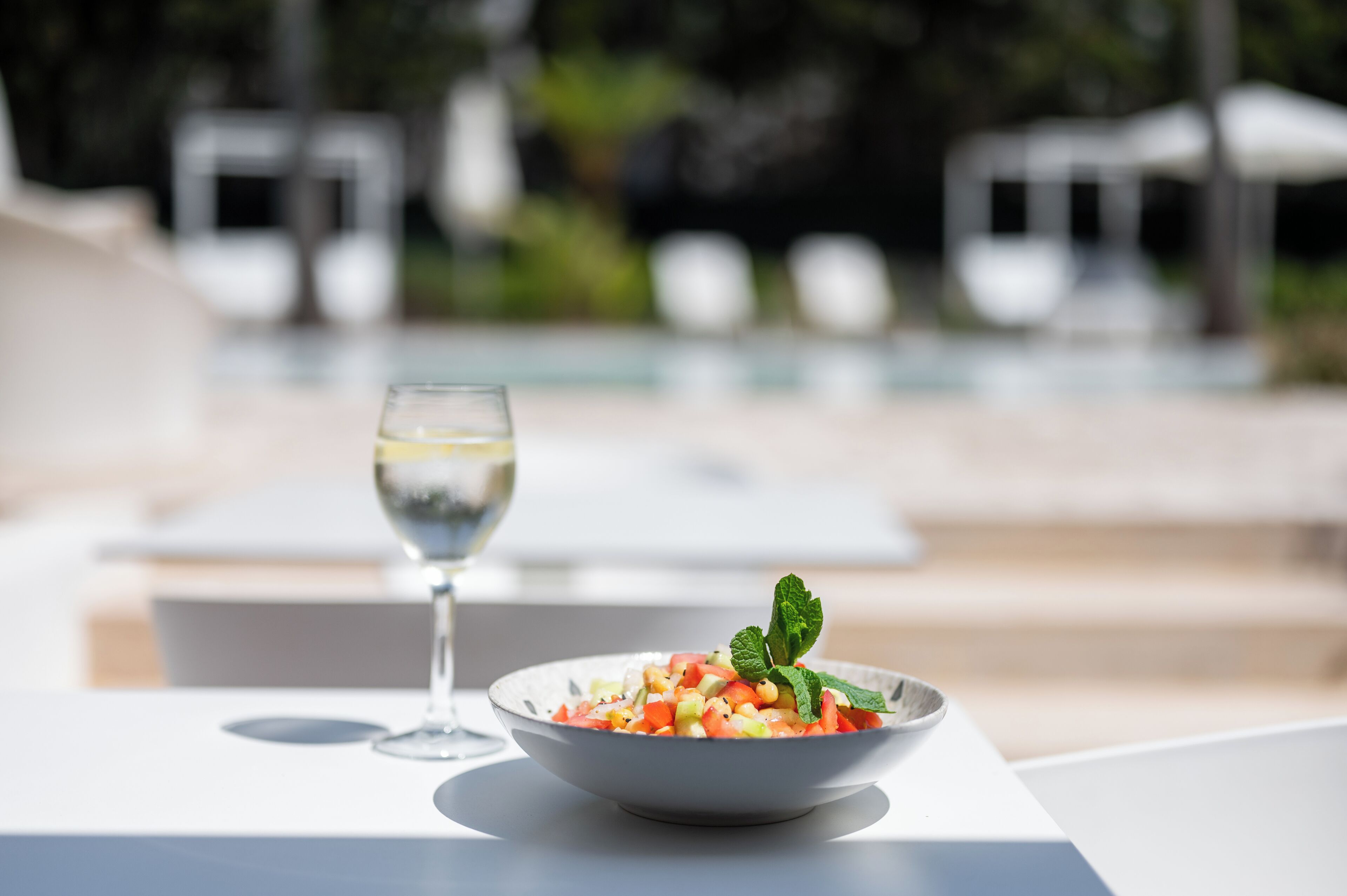 Un saladier rempli de salade de fruits, garni de feuilles de menthe, est posé sur une table blanche au bord d’une piscine.
