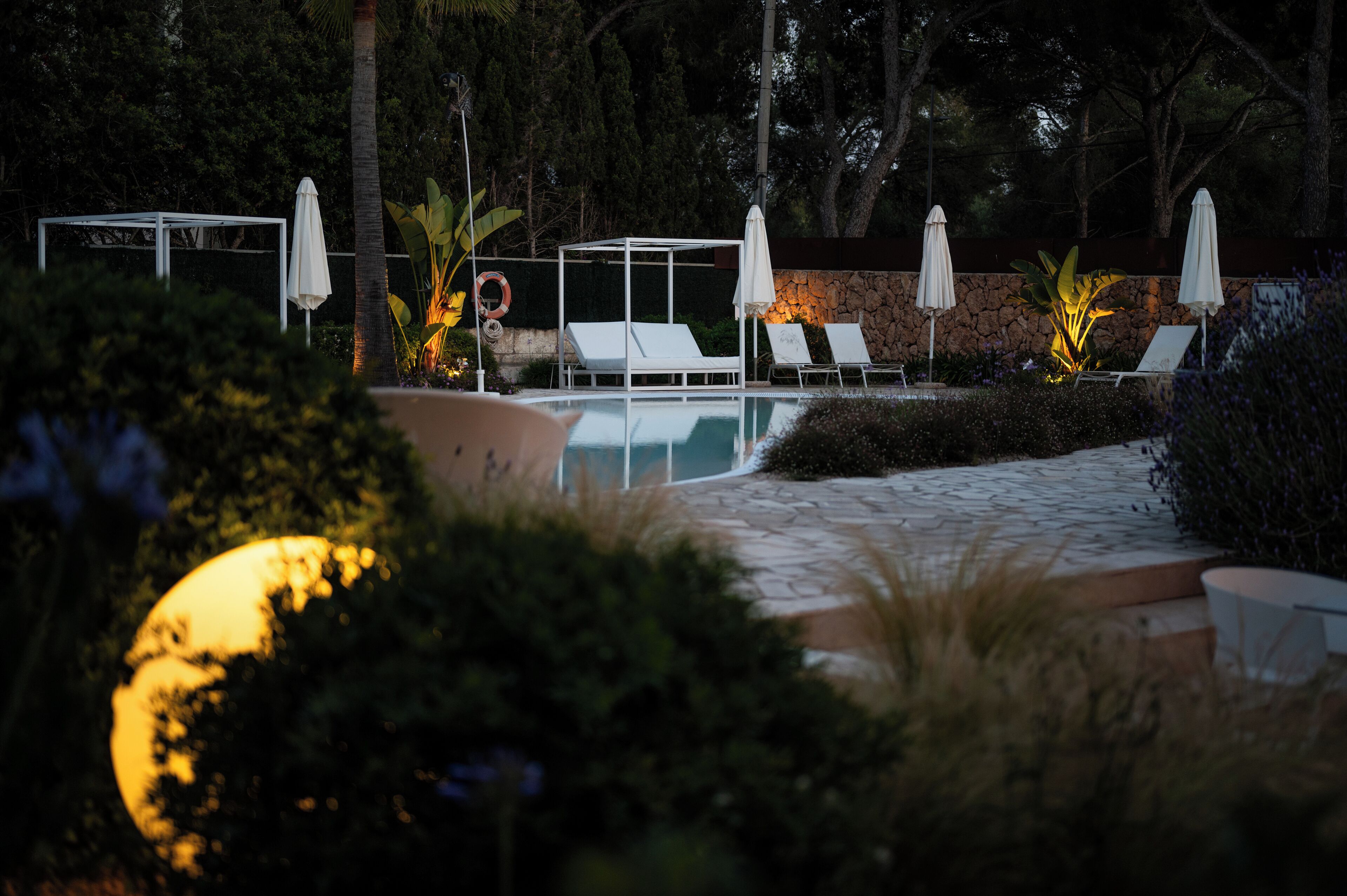 A pool area with lounge chairs and umbrellas, surrounded by greenery and a stone wall.