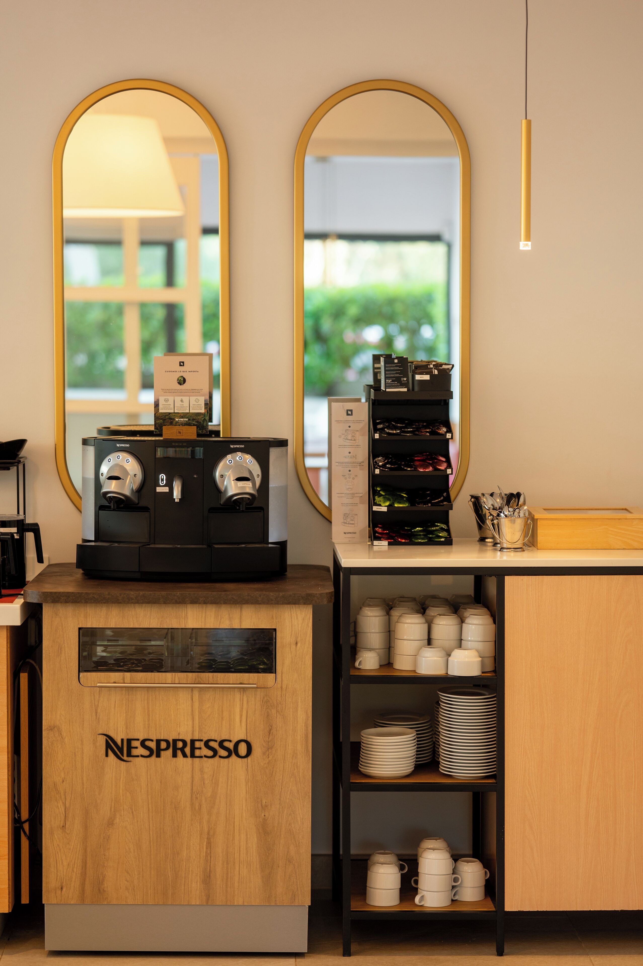 Una estación de café Nespresso con una cafetera, una pila de cápsulas de café y una repisa con tazas y platos.