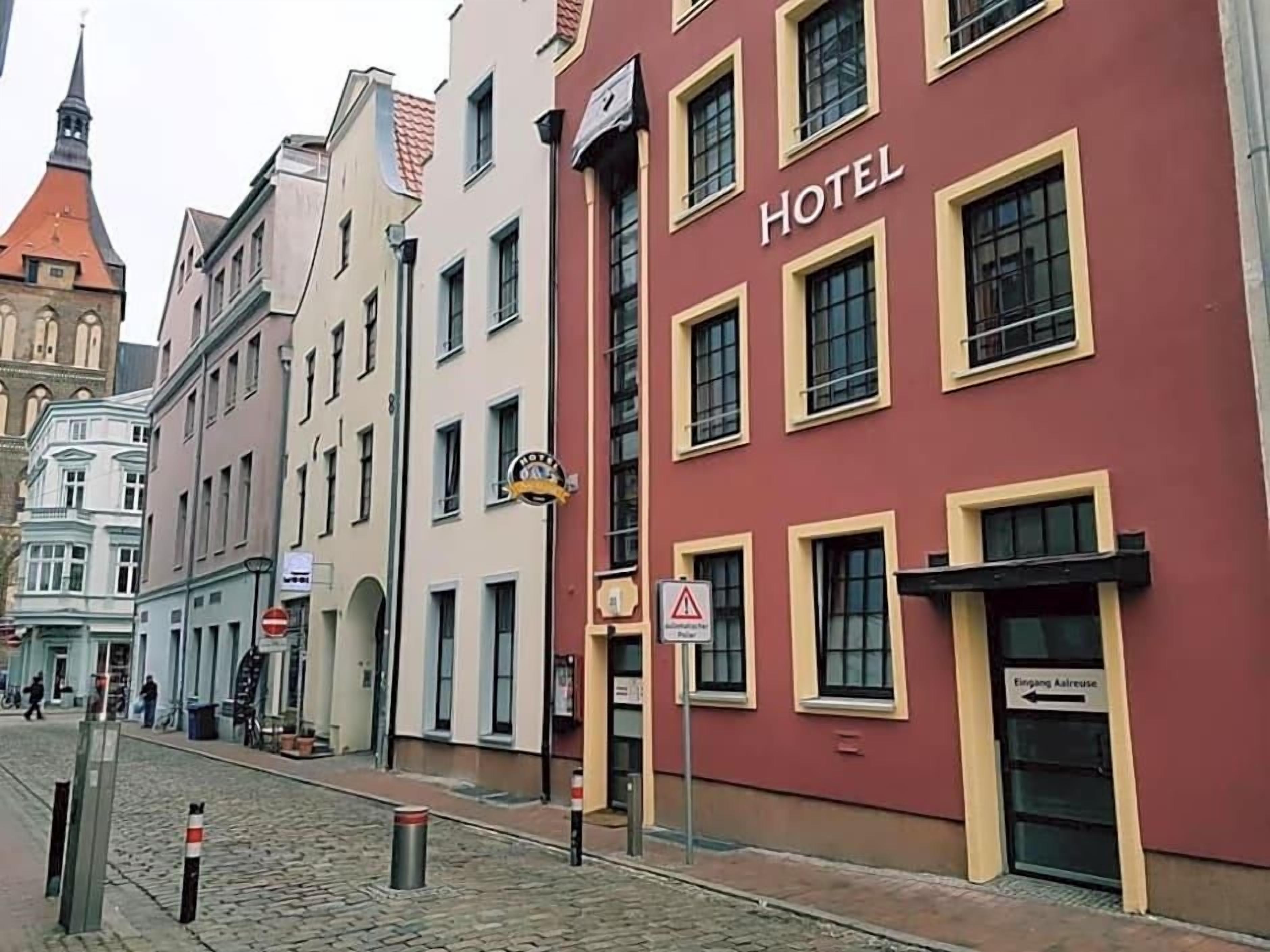 Ein Hotelgebäude mit roter Fassade und gelben Fensterrahmen, eine Kopfsteinpflasterstraße sowie ein Kirchturm im Hintergrund.