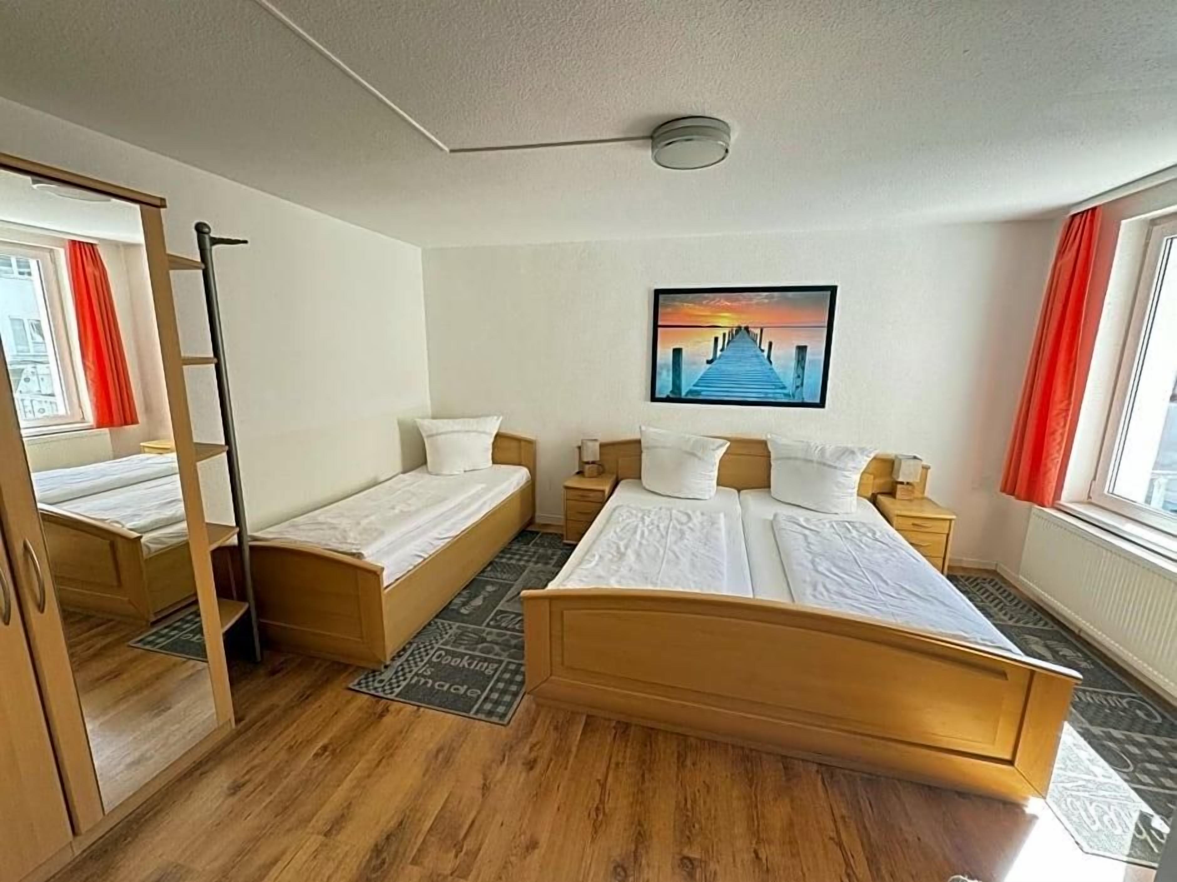 A hotel room with a large bed, two smaller beds, a wardrobe, a painting, and a window with curtains.