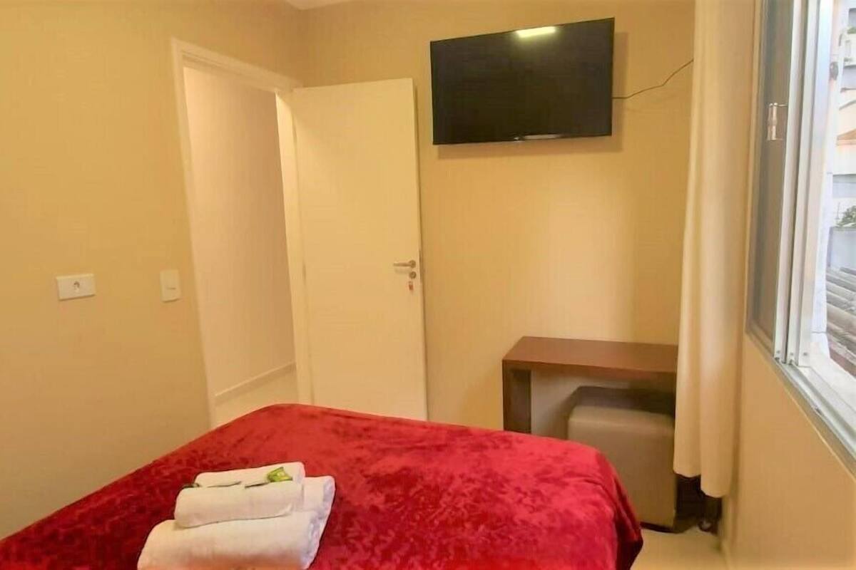 A hotel room with a bed, a TV mounted on the wall, a small table, and a window with curtains.