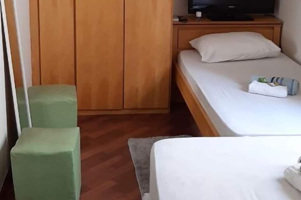 A bedroom with a wooden wardrobe, a bed with white bedding, a green chair, and a television on a wooden nightstand.
