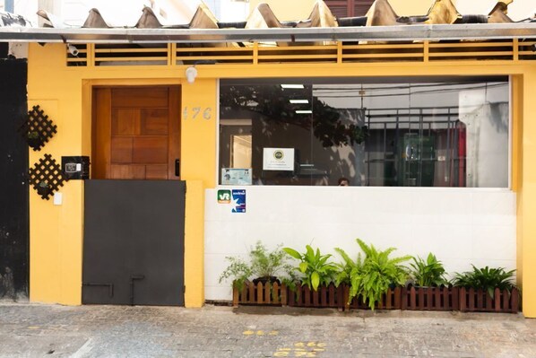 A yellow building with a wooden door marked '176', a window with a sign, and a small fence with plants.