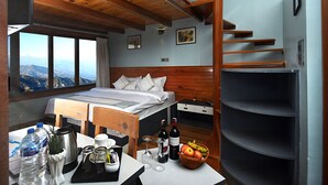Ein Schlafzimmer mit Bett, Holz-Kopfteil, Schreibtisch und Obstkorb sowie Bergblick durch große Fenster.