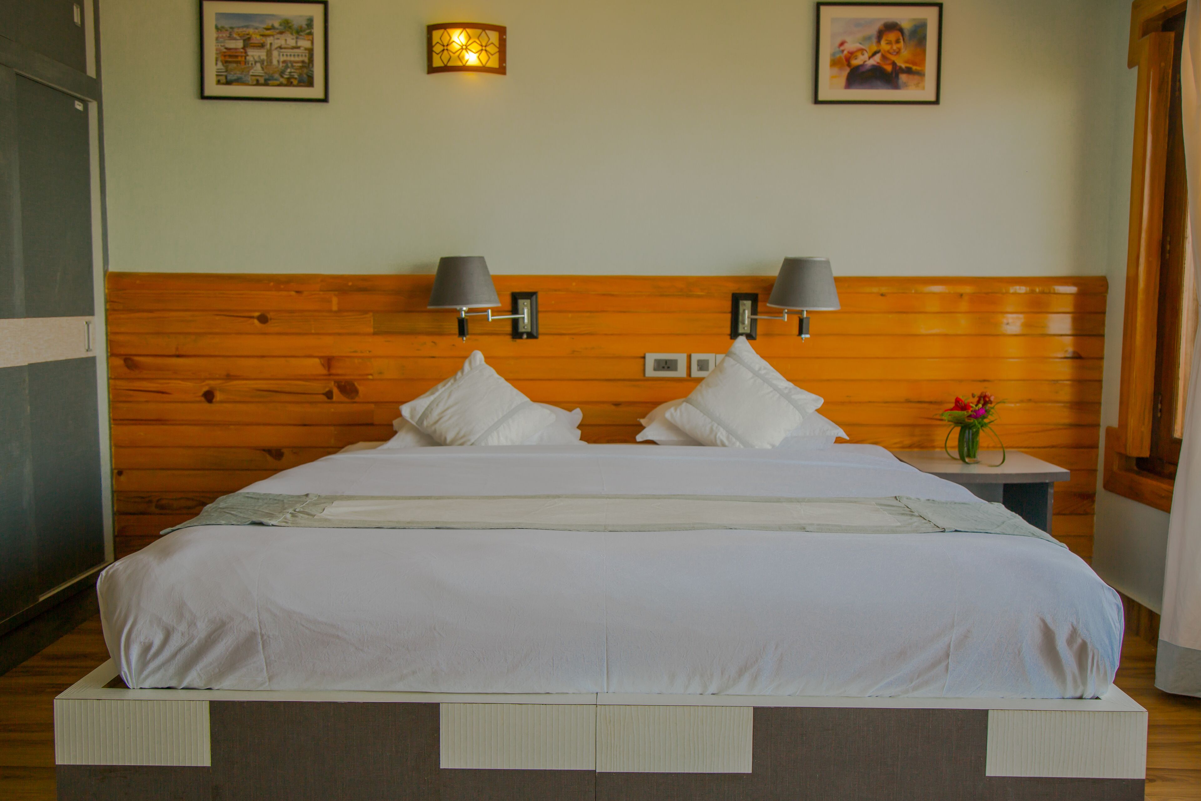 A neatly made bed with white linens and pillows, a wooden headboard, two wall-mounted lamps, and a small table with a vase of flowers.