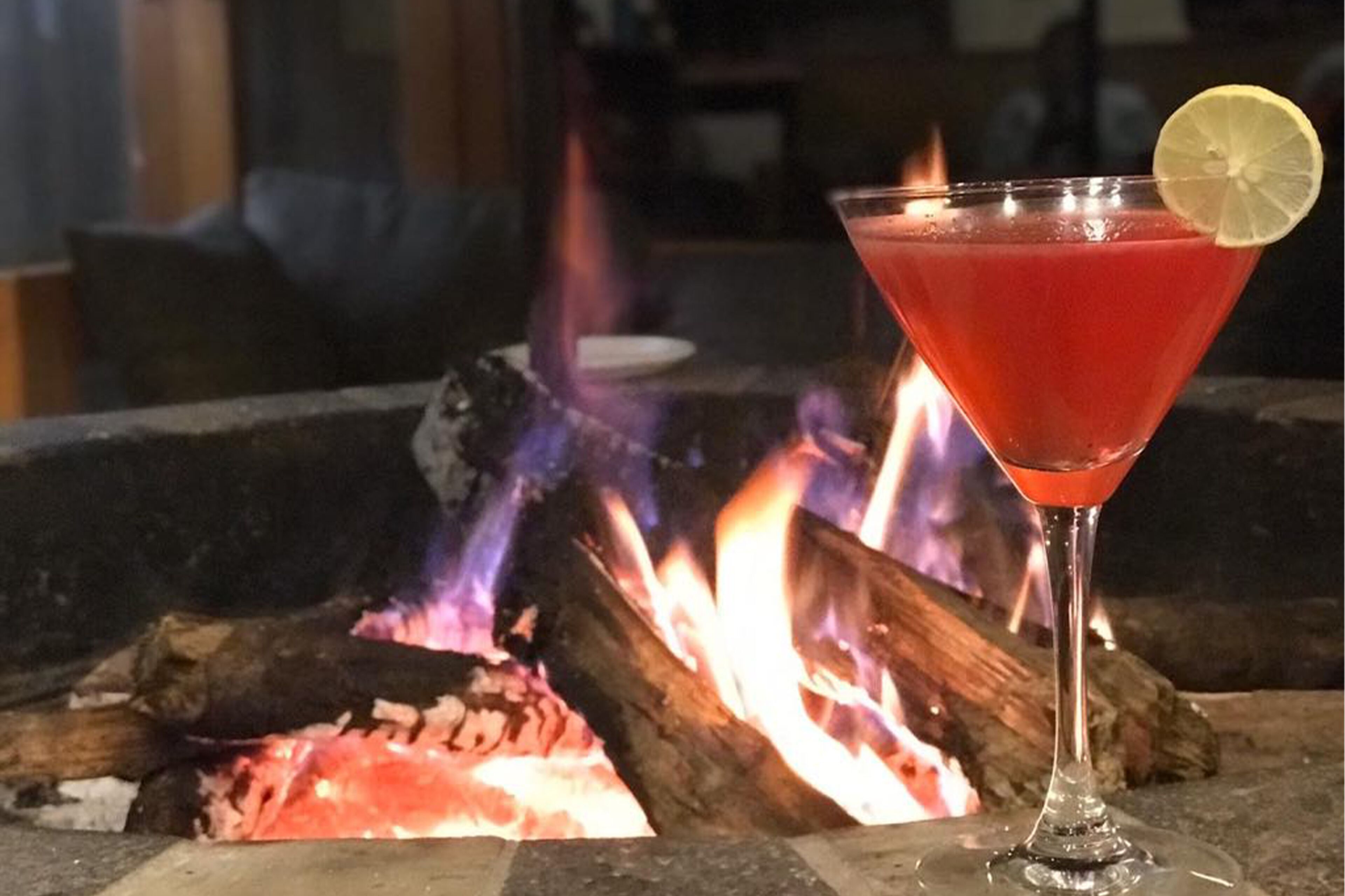 Un cóctel en una copa de martini con adorno de limón, colocado frente a un hogar.