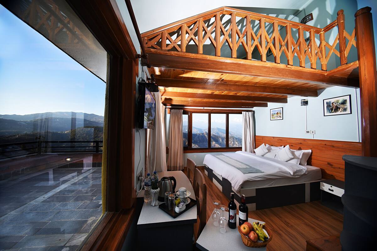 A bedroom with a bed, a dining table with breakfast items, a view of the mountains, and a balcony with a wooden railing.