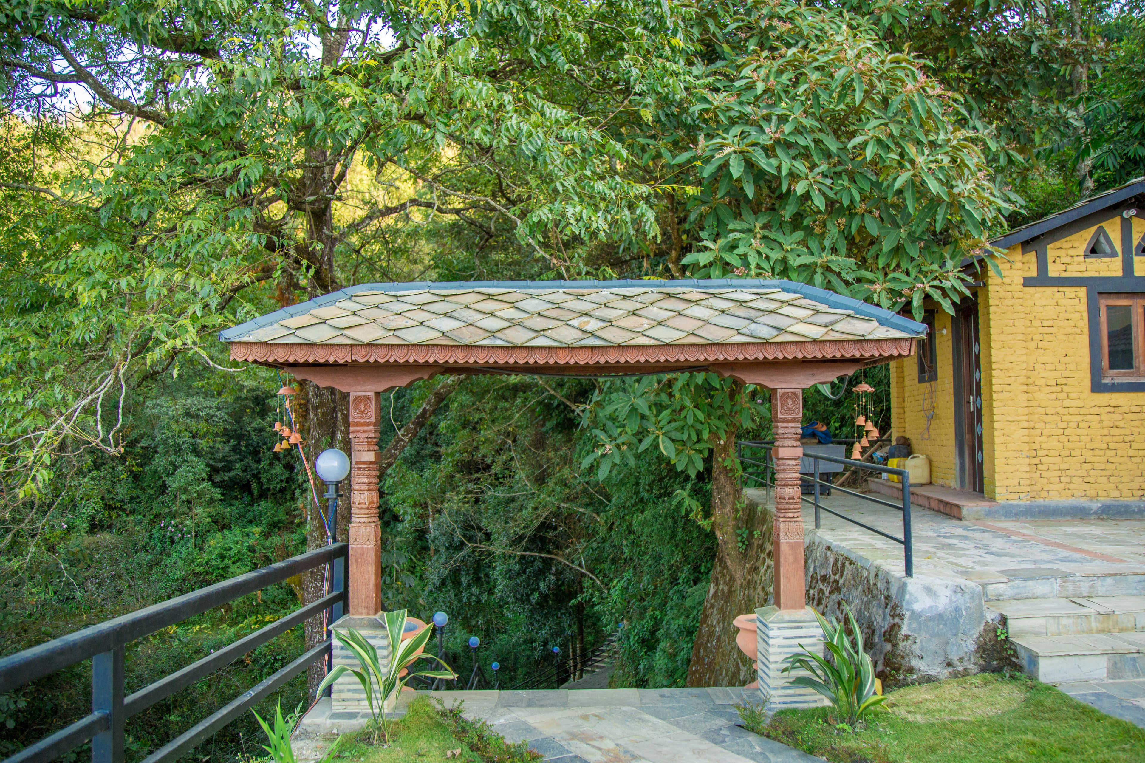 A gazebo with a tiled roof, surrounded by lush greenery and a stone pathway leading to a building with a yellow facade.