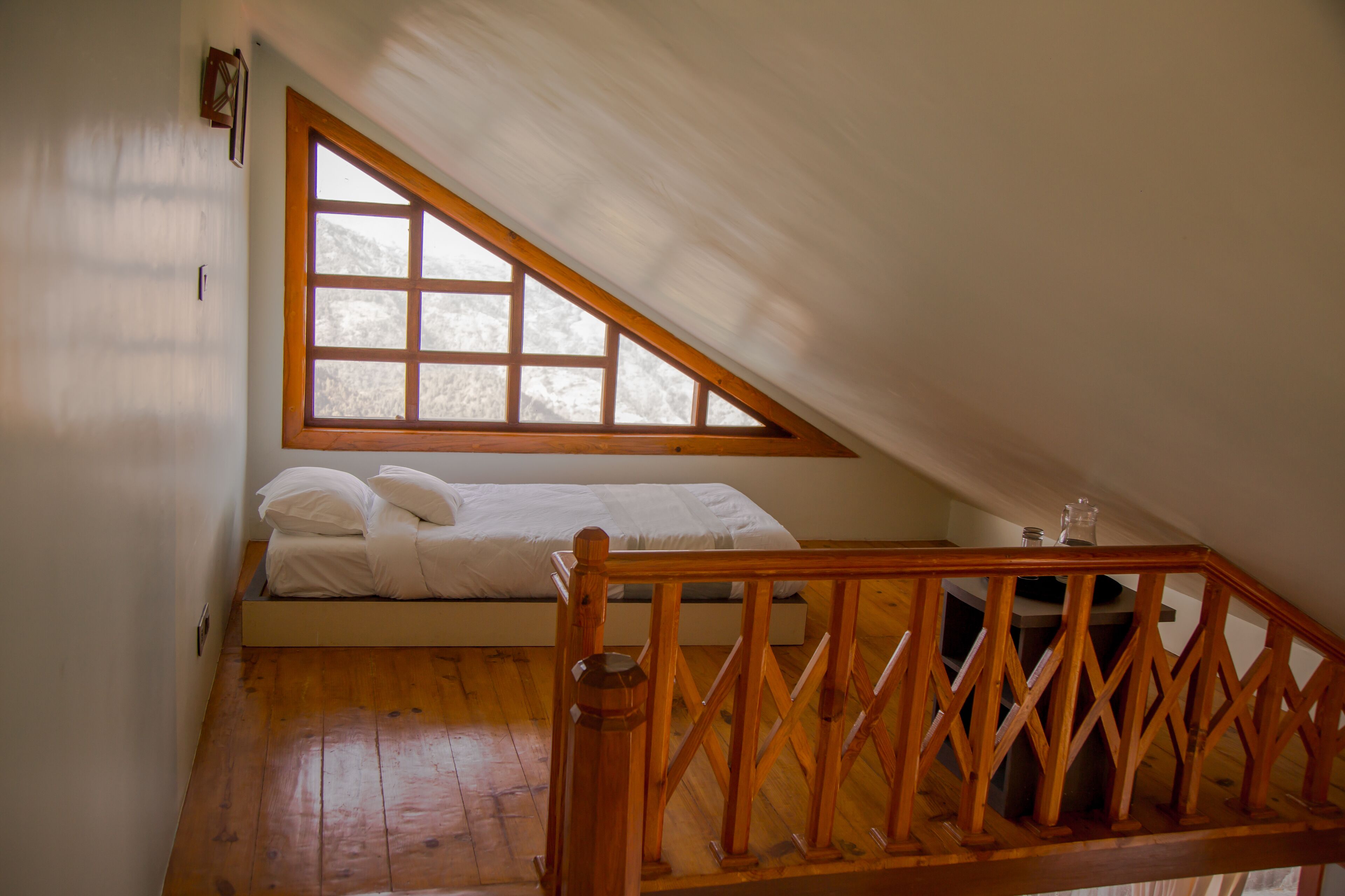 Une chambre avec un plancher en bois, un lit recouvert de draps blancs, une fenêtre avec un cadre en bois et une balustrade en bois.