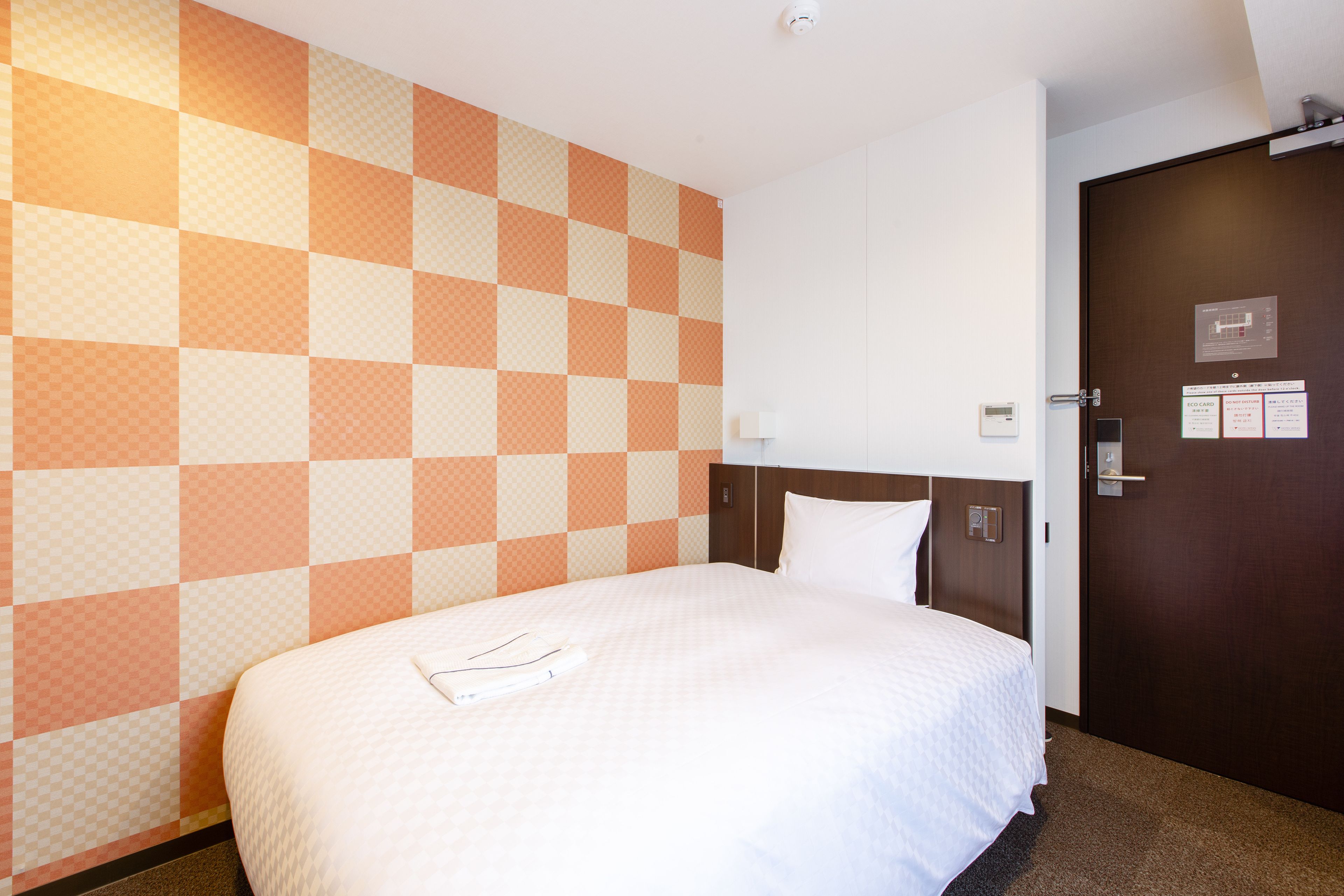 A hotel room with a checkered wall pattern, a neatly made bed, a wooden headboard, and a door with a lock and a light switch.