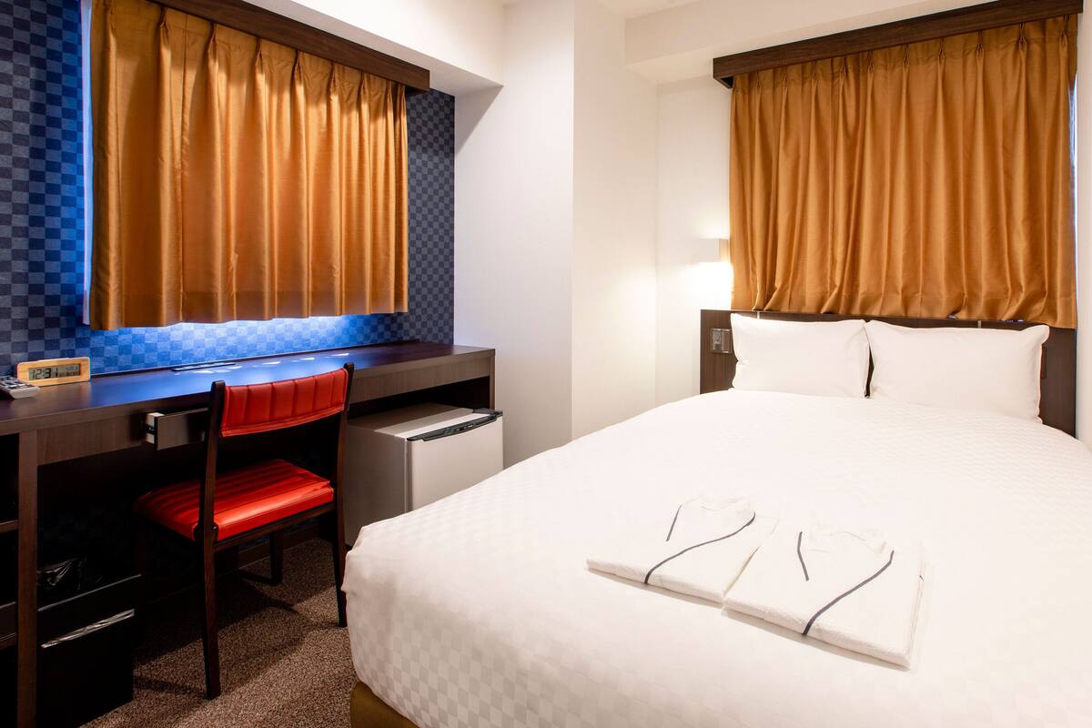 Une chambre d’hôtel comprenant un lit, un bureau avec une chaise, un petit réfrigérateur et une fenêtre avec des rideaux.