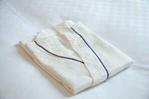 A neatly folded white bathrobe with a dark trim on a bed with a patterned white sheet. - KOKO HOTEL Akabane Station (Tokyo)