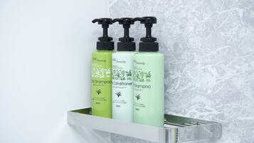 Três frascos de shampoo e condicionador DHC Amenity sobre uma prateleira branca, em frente a uma parede de mármore.