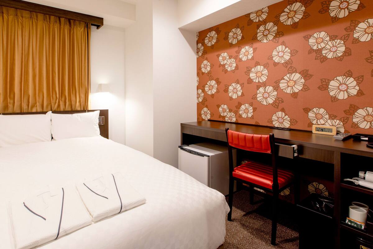 Une chambre d’hôtel comprenant un lit, un bureau avec une chaise, une horloge et un mur orné d’un motif floral.