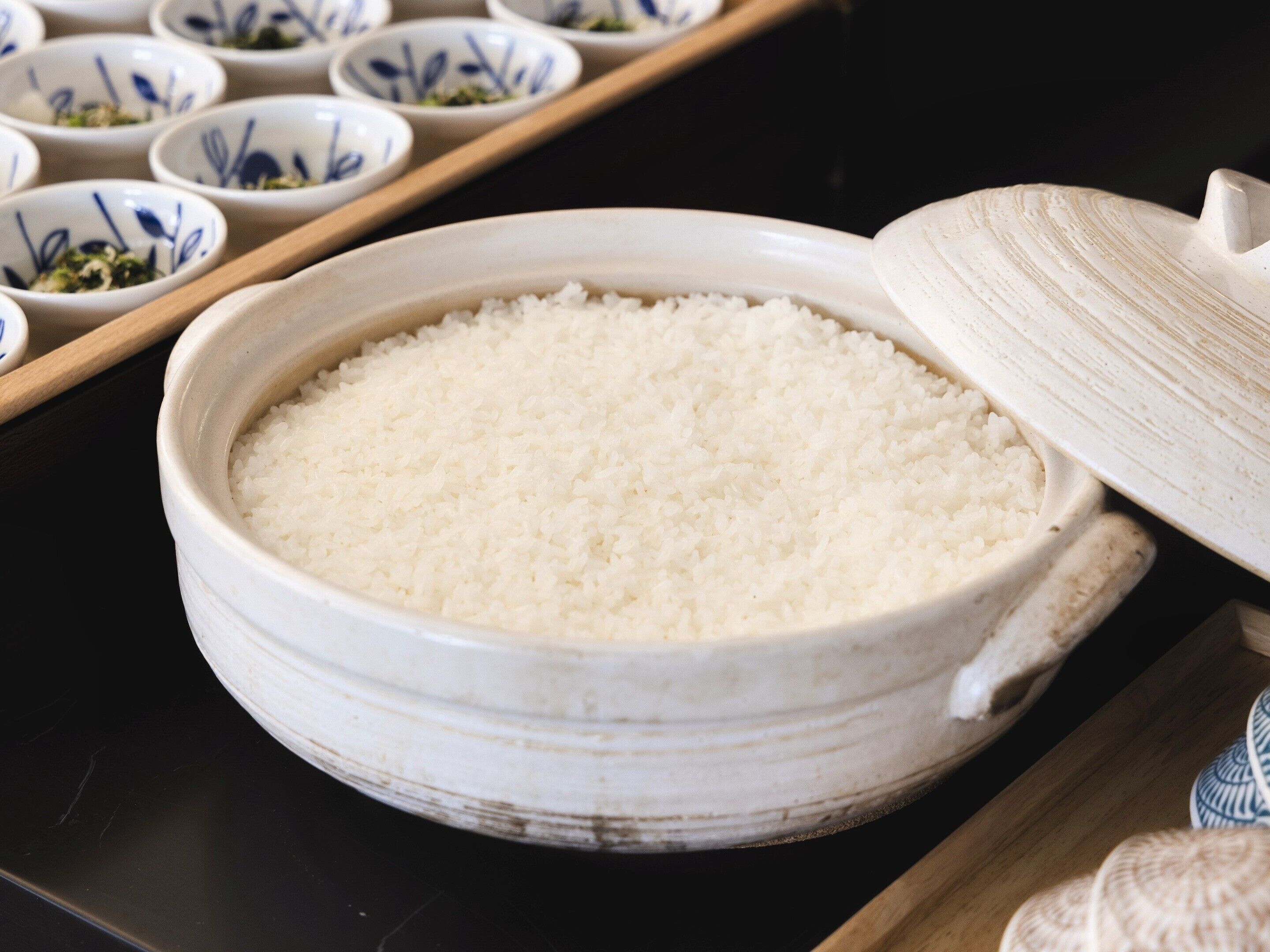 A large ceramic bowl filled with white rice, placed on a dark surface with a wooden tray containing smaller bowls in the background.
