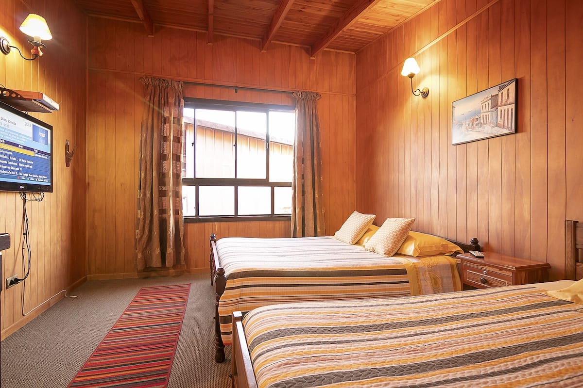 A wooden-paneled room with two beds, a window with curtains, a television, and a framed picture on the wall.