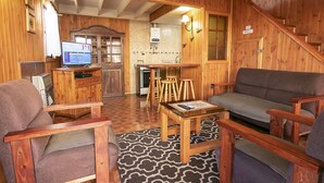 Un interior de madera acogedor con una sala de estar que incluye un sofá, sillones y una mesa de centro. Un televisor está montado en un mueble de madera, y se ve un área de cocina al fondo.