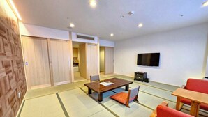 A traditional Japanese-style room with tatami flooring, a low table with cushions, a television mounted on the wall, and sliding doors leading to a kitchen area. - Santoka - Kurashiki Yuga Onsen - (Kurashiki)