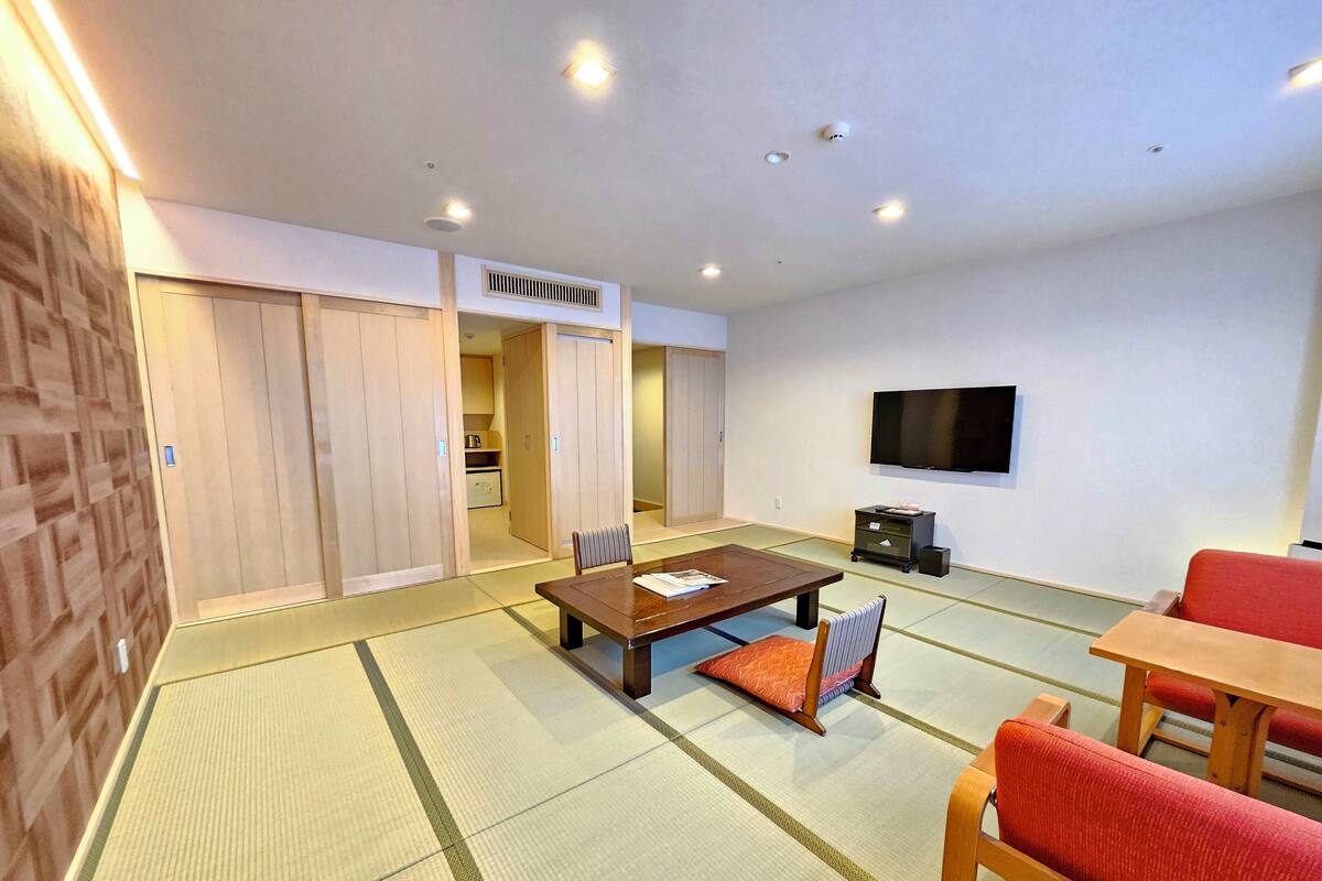Una habitación de estilo japonés tradicional con suelo de tatami, una mesa baja con cojines, un televisor montado en la pared y puertas correderas que dan a una zona de cocina.