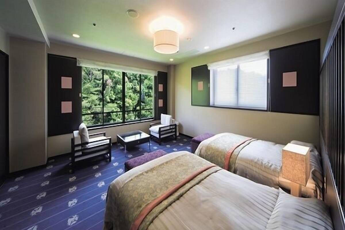 Habitación de hotel con dos camas, un sofá, una mesita y ventana con vistas a zonas verdes.