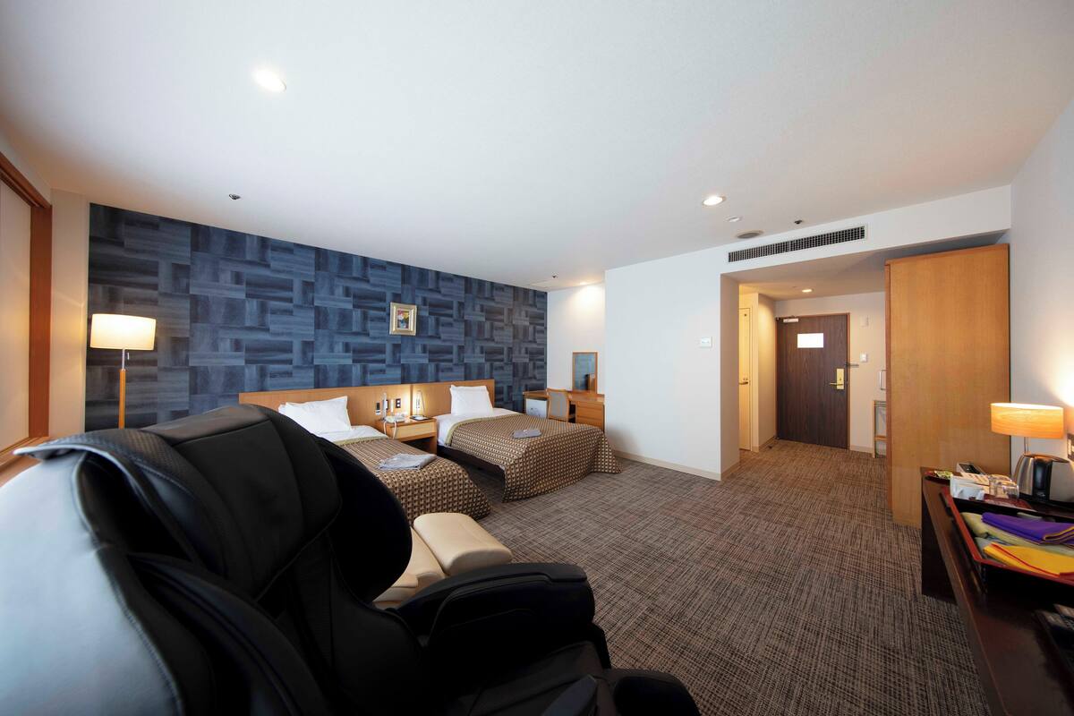 Una habitación de hotel con cama, dos mesitas de noche, un escritorio con silla, un sillón de masaje, una pared con diseño geométrico y una puerta que da a otra habitación.