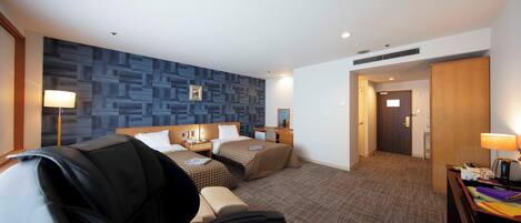 Una habitación de hotel con cama, dos mesitas de noche, un escritorio con silla, un sillón de masaje, una pared con diseño geométrico y una puerta que da a otra habitación.