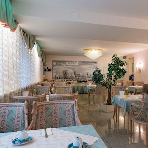 A dining area with tables set for tea service, chairs with patterned upholstery, a large cityscape painting on the wall, and a chandelier. - Hotel Villa Sulmana (Neckarsulm)