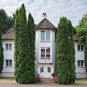 A two-story building with a grey facade, a weather vane on the roof, and a symmetrical window arrangement.