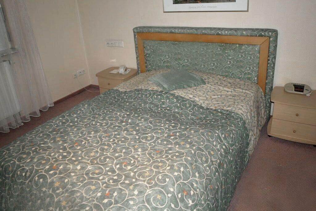 Ein ordentlich bezogenes Bett mit einem gemusterten Überwurf, ein Nachttisch mit einer Lampe und ein kleiner Nachttisch mit einer Uhr.