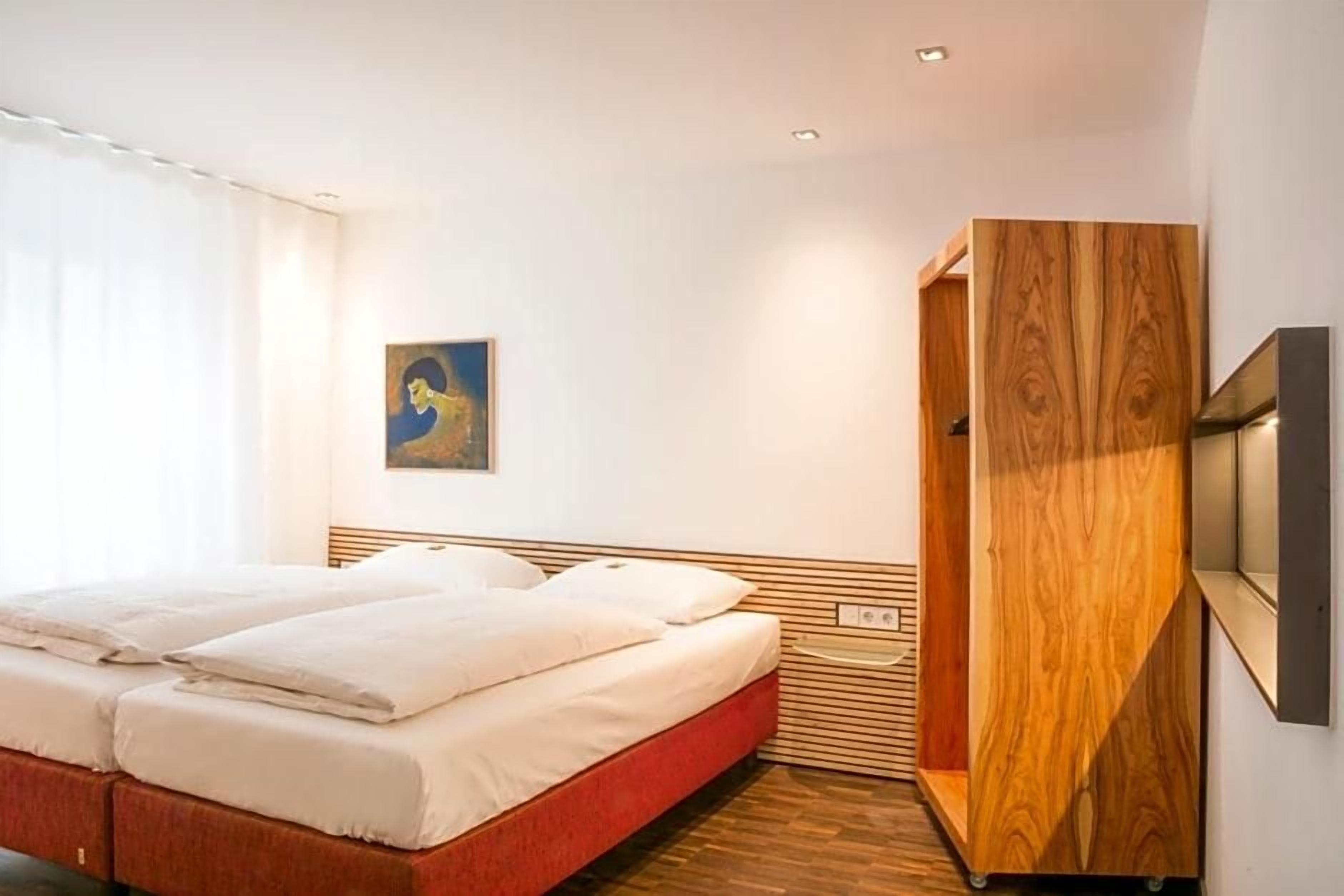Una habitación de hotel con una cama, un armario de madera, un cuadro en la pared y una ventana con cortinas.