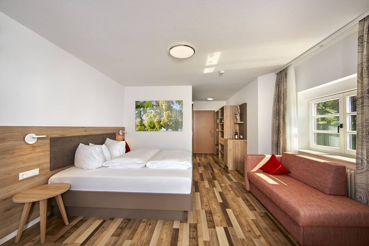 A modern hotel room with a bed, a small table, a sofa, a wooden floor, and a window with curtains.
