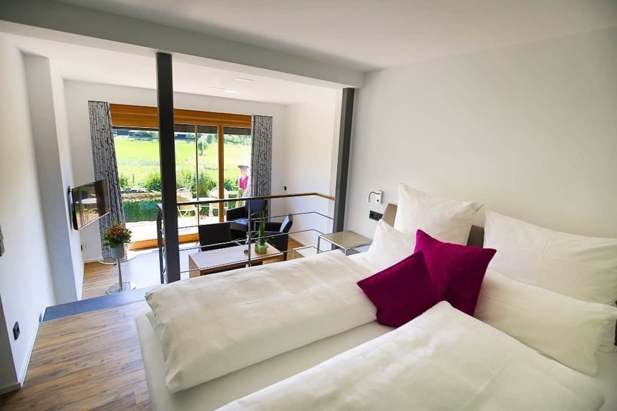 A modern hotel room with a balcony, a bed with white and pink pillows, and a view of a green landscape.